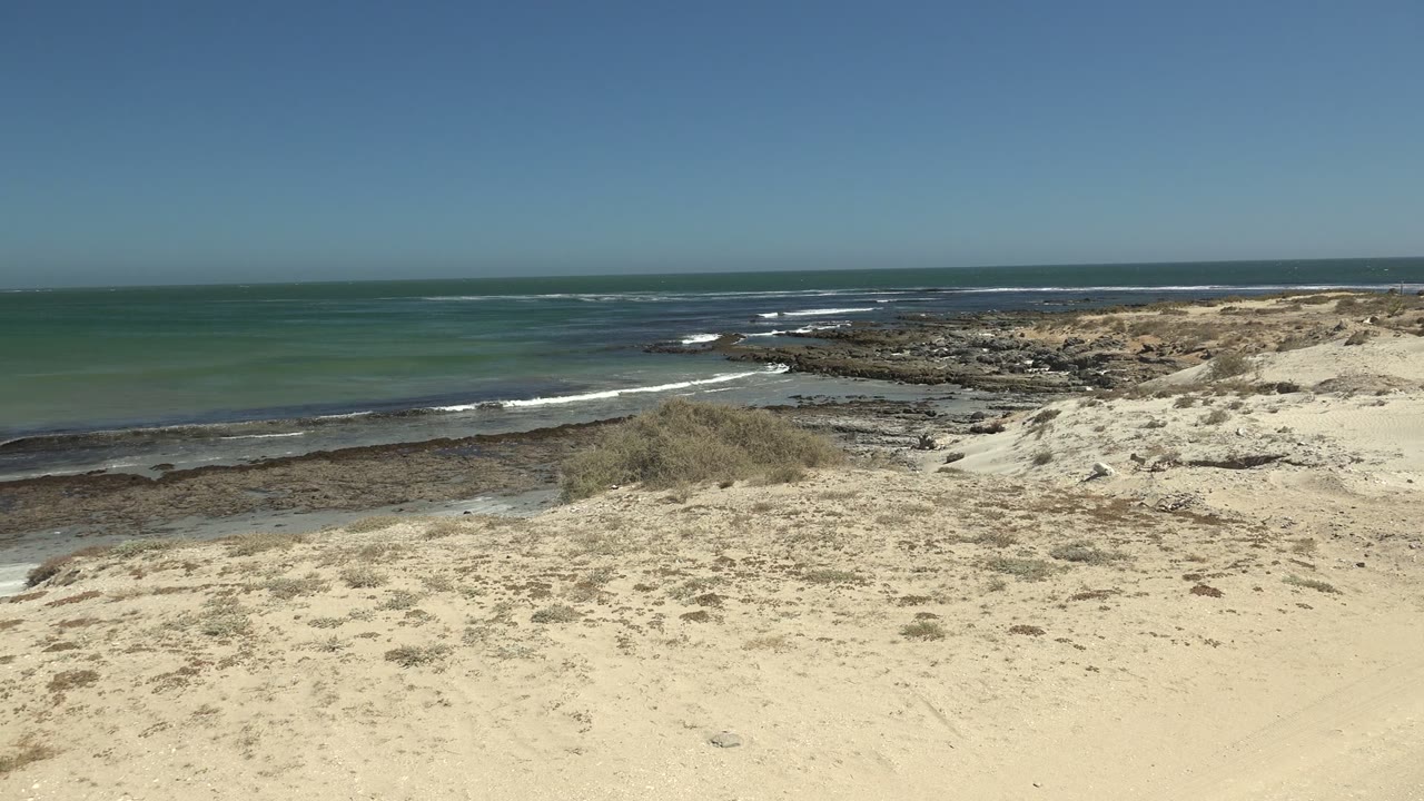 At 0:00, La Bocana sand and sparse brush fill the foreground above a rocky shoreline, green-blue ocean, low white waves, clear sky, and no visible people.