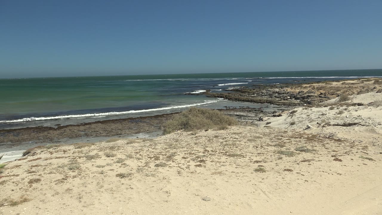 Open beach sand with rocky shore, ocean waves, and clear sky.
