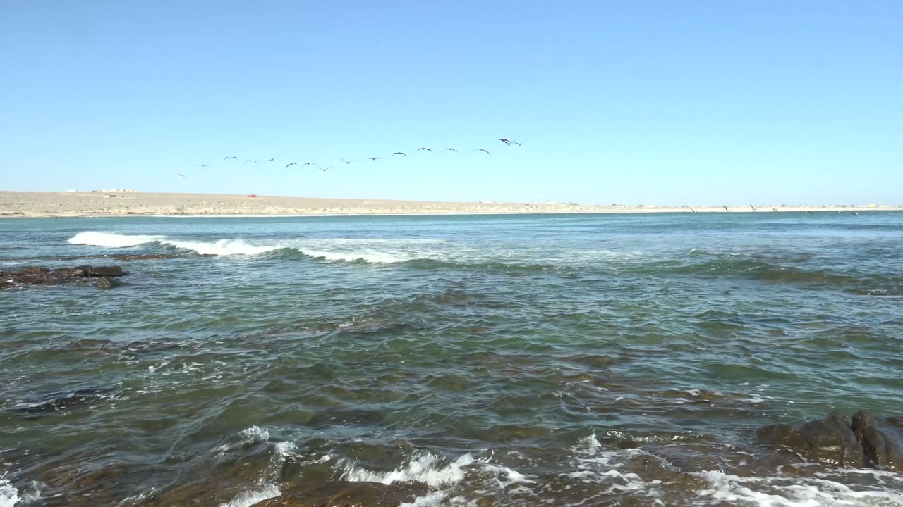 At La Bocana in Baja California Sur, the camera view holds on a broad stretch of ocean with foamy wave lines, dark rocks, distant low coastline, and small birds in the sky, with no visible people.