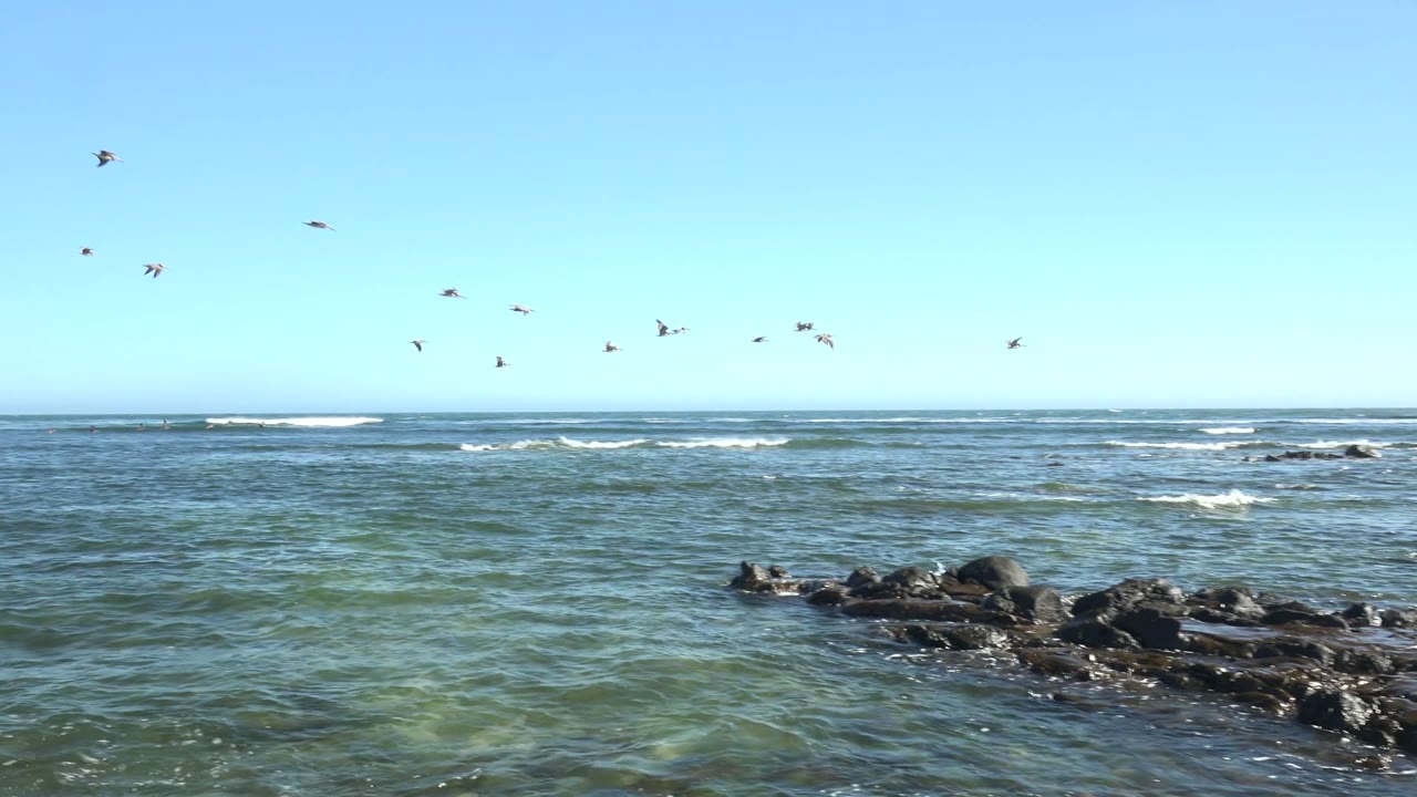 The rocky shore at La Bocana occupies more of the foreground, with shallow green-blue water moving around the rocks, seabirds scattered near the horizon, and no visible people.