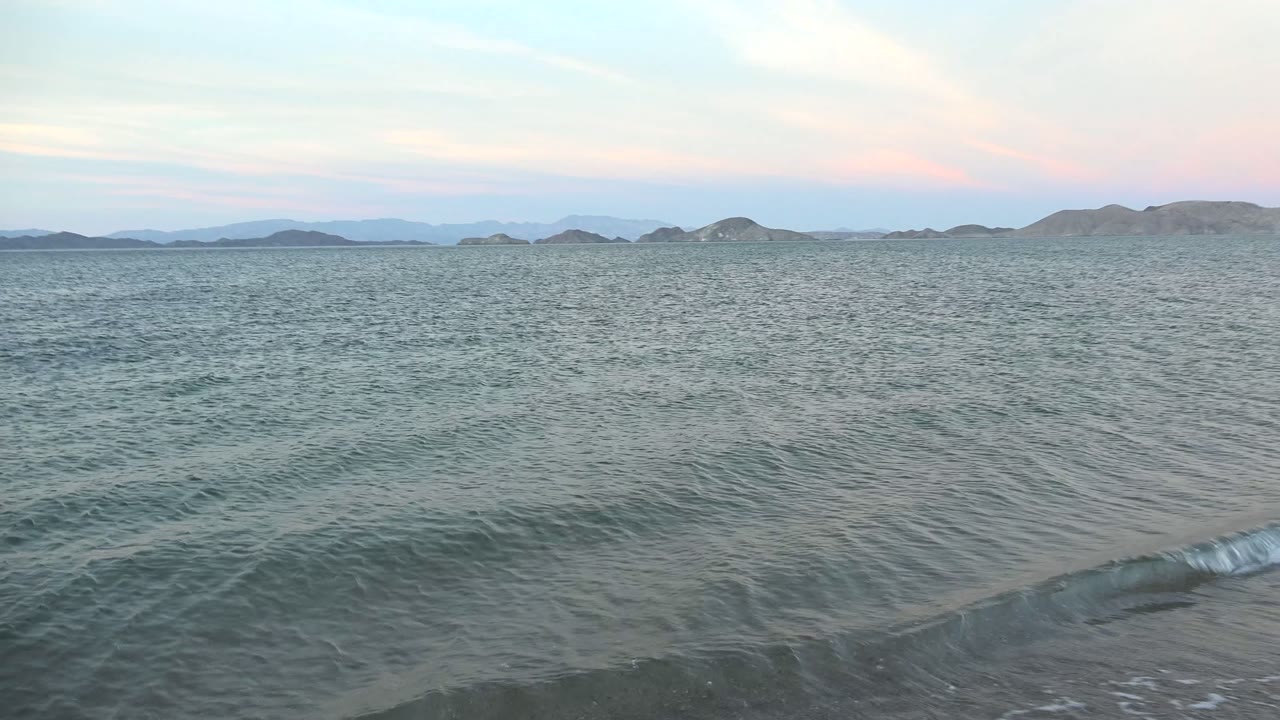 At Bahia De Los Angeles, small waves roll toward wet sand with distant low hills on the horizon and no visible people.