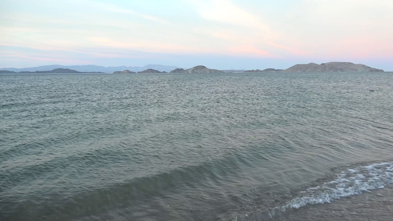 Low ocean swell forming near the beach with islands or hills beyond.