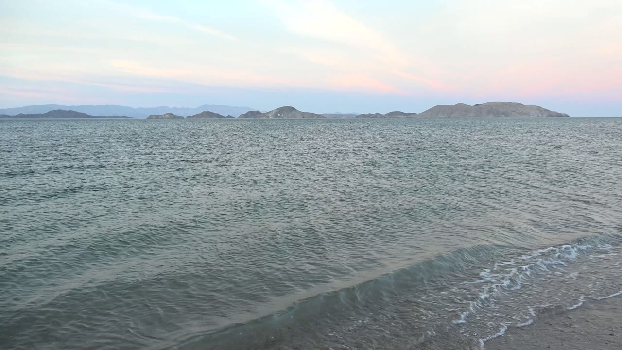 Foam curls along the right side of the Bahia De Los Angeles shoreline while open water stretches to low hills beneath pale dusk clouds, no visible people.