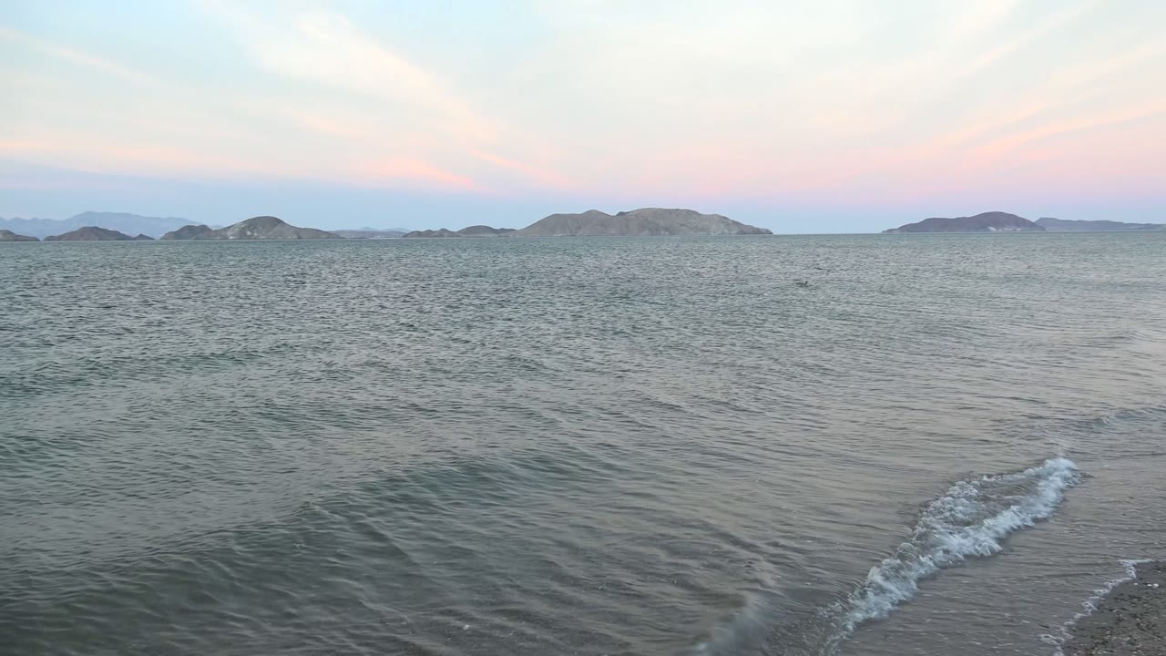 Wide Bahia De Los Angeles shoreline view with small waves in the foreground, low islands on the horizon, pastel dusk sky, and no visible people.