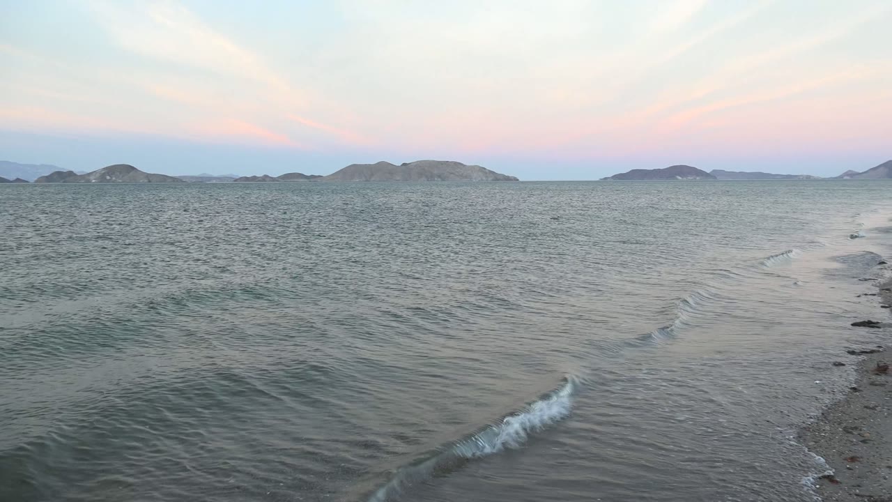 Ground-level Bahia De Los Angeles shoreline composition with foamy water tracking along the right side, textured sea to the left, island silhouettes ahead, and no visible people.