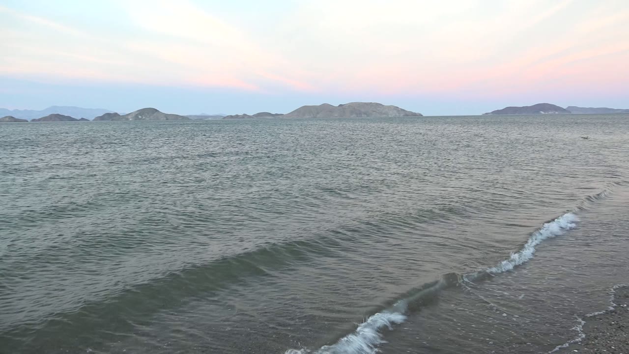 Wide view of gentle waves reaching a sandy Bahia De Los Angeles shoreline under a pale pink and blue dusk sky, with low hills across the horizon and no visible people.