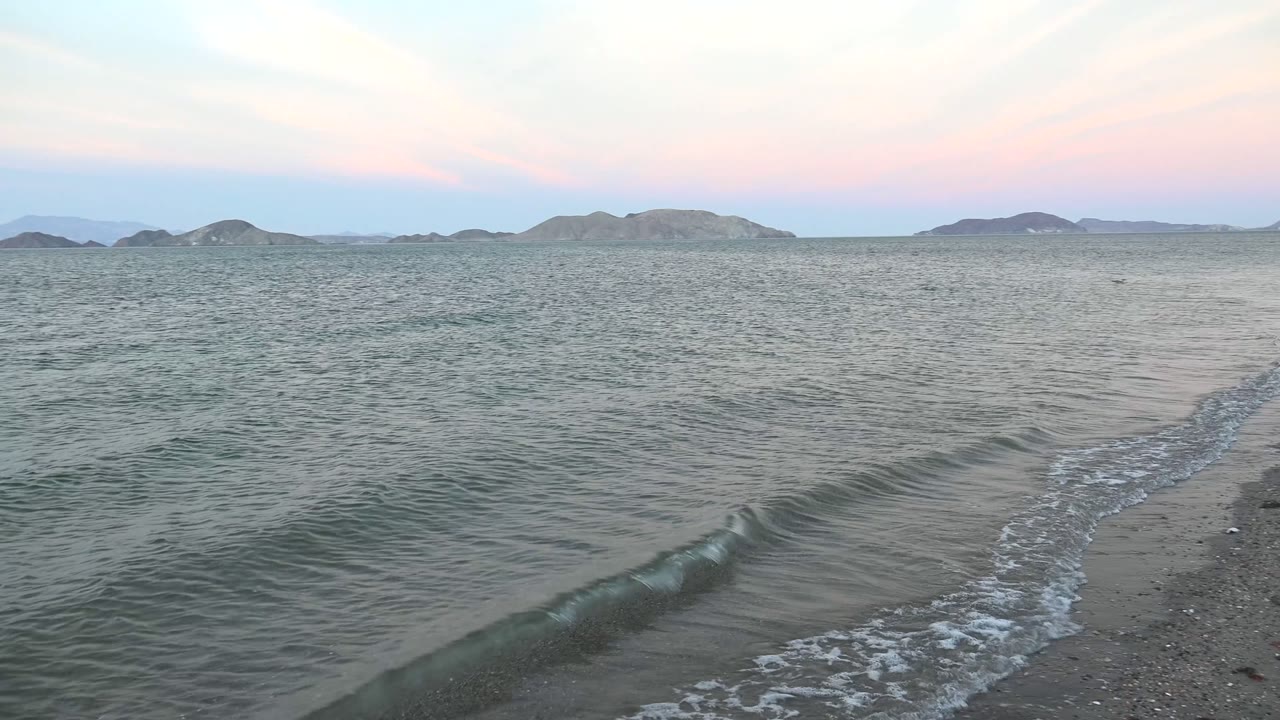 A small wave breaks near the beach with distant hills beyond the water.