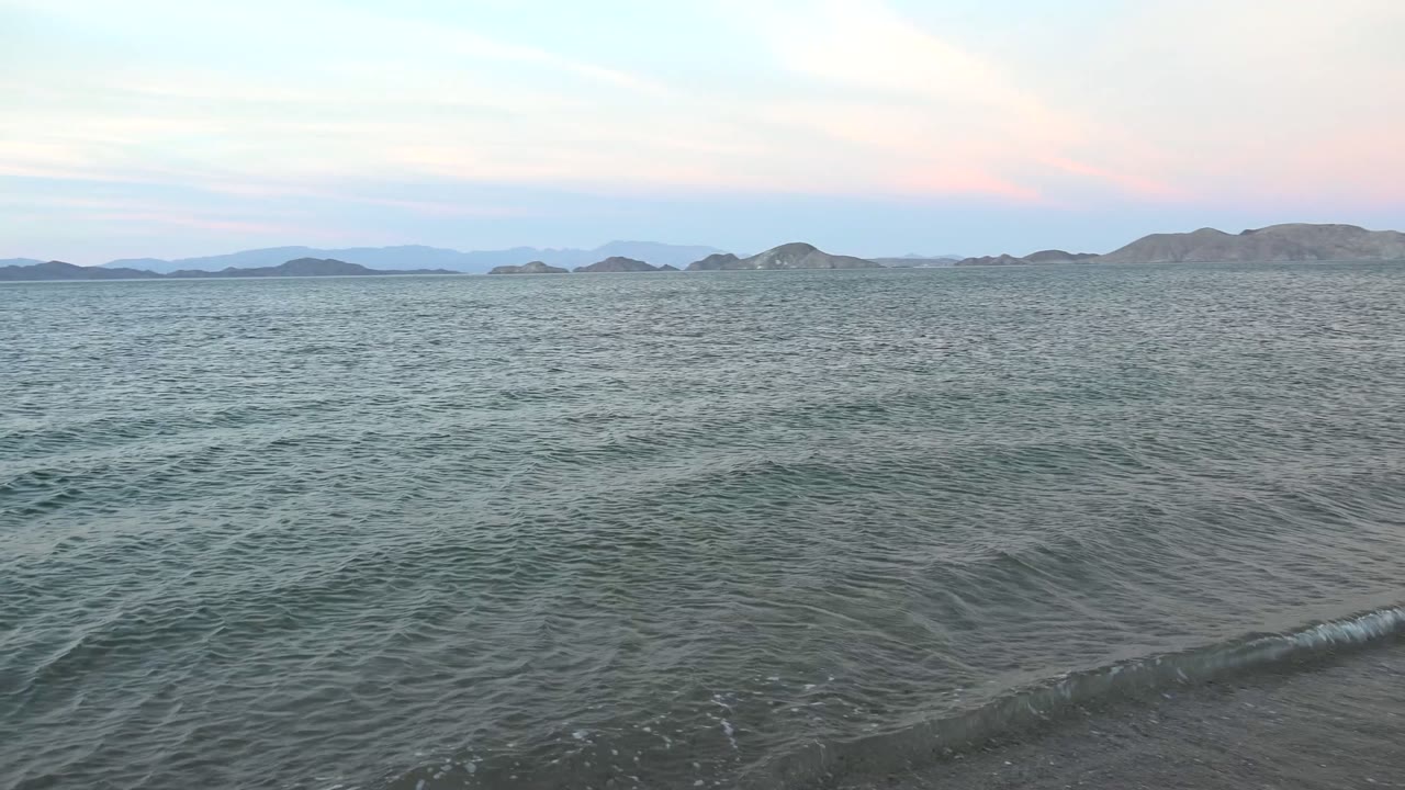 Small wave line approaching a sandy shoreline with hills in the distance.