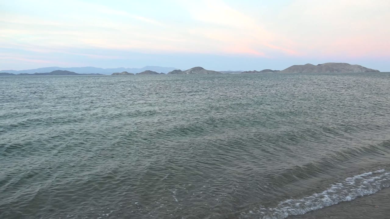 A small crest breaks near the sandy edge in Bahia De Los Angeles while gray-green water fills the foreground, low hills span the horizon, and no visible people appear in the frame.