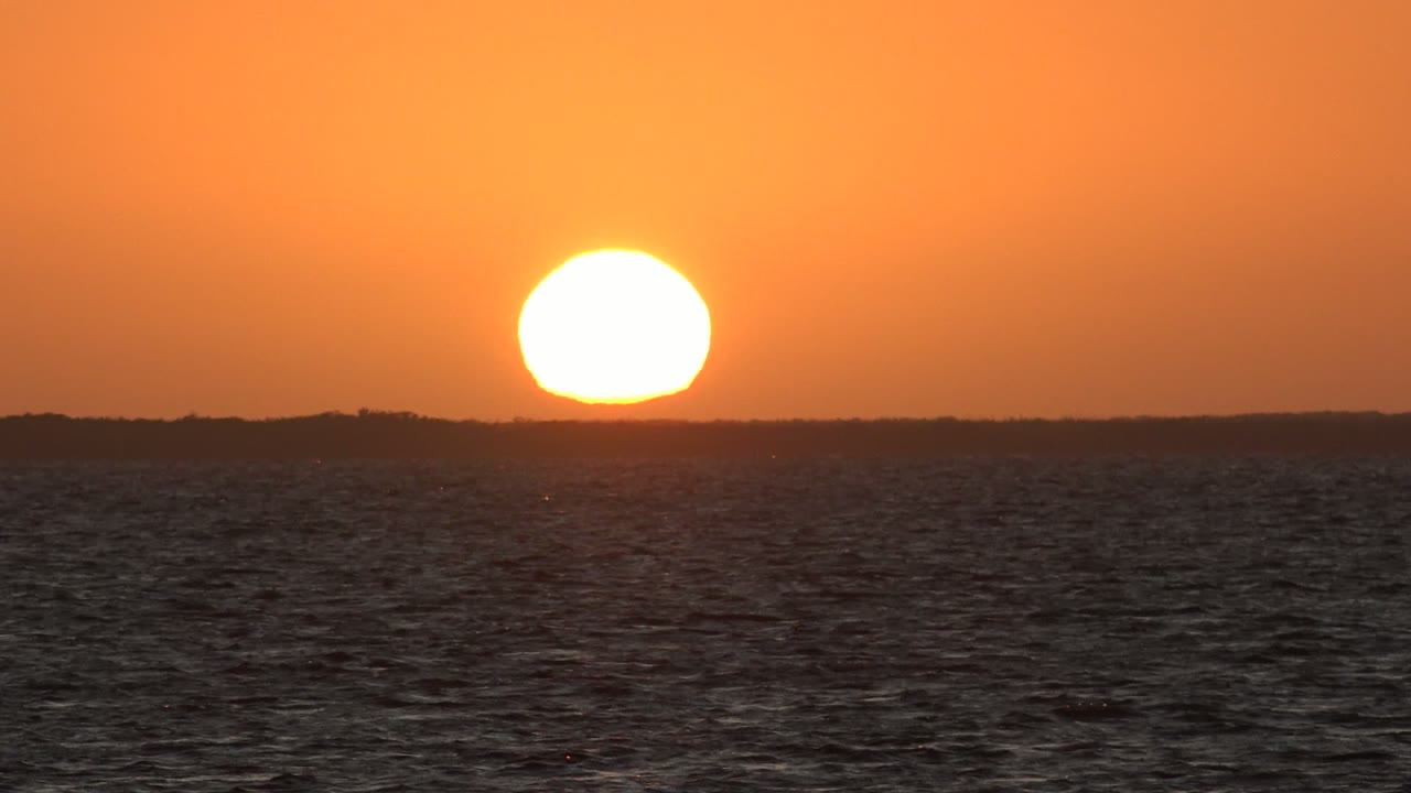 At 0:00 in San Carlos, Baja California Sur, the bright sun sits just above the dark ocean horizon with rippled water in the foreground and no visible people.