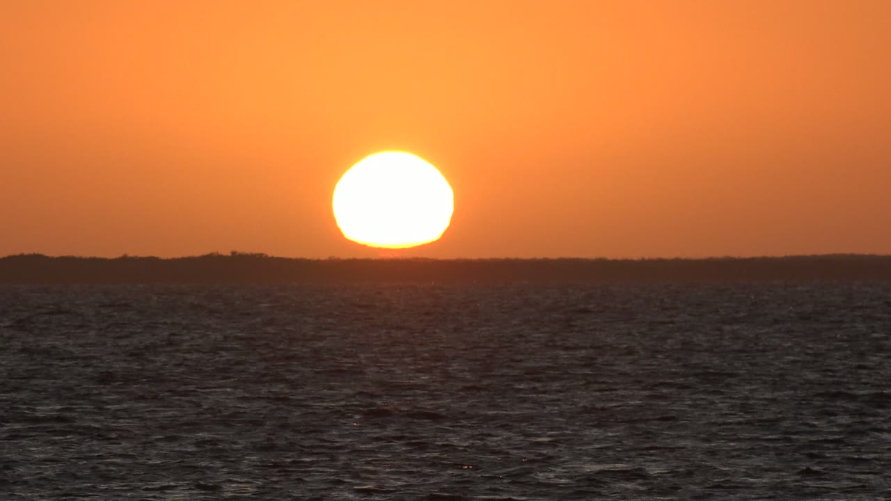 Bright sun over rippled ocean with a low land horizon.