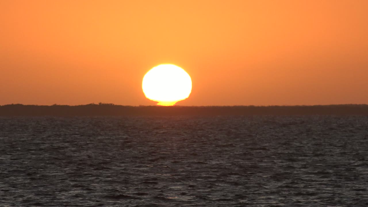 Low orange sun hangs just above the distant San Carlos, Baja California Sur shoreline, with rippled ocean in the foreground and no visible people.