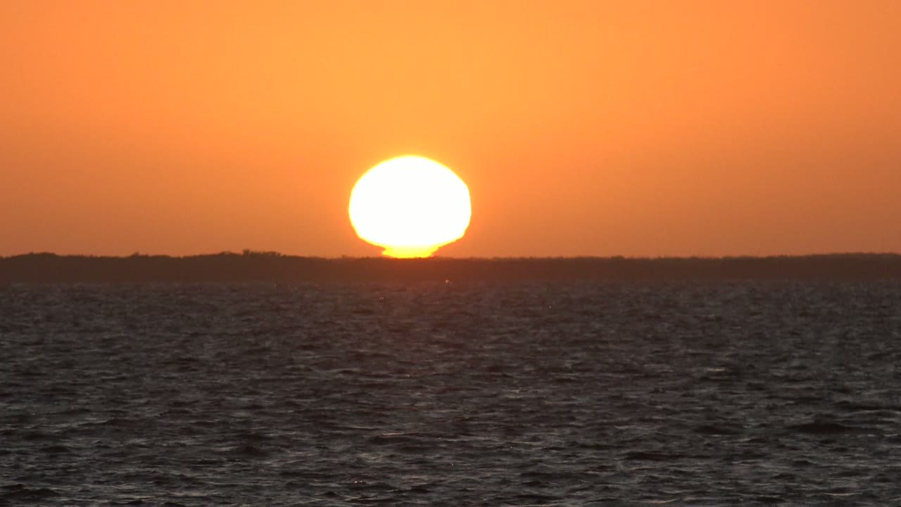 A large white-yellow sun sits over a thin dark land silhouette in San Carlos, Baja California Sur, while the ocean surface fills the lower half and no people are visible.