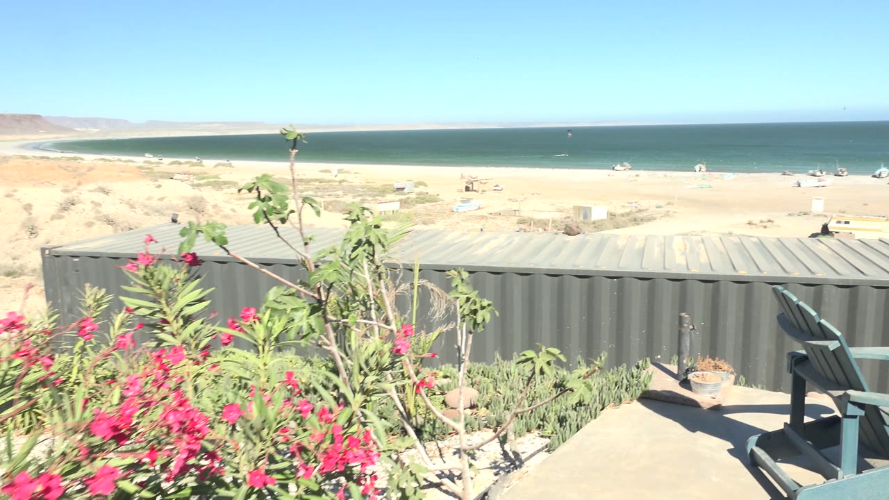 Elevated view in San Juanico, Baja California Sur showing pink flowers, chairs, a gray metal foreground, sandy beach, blue ocean, clear sky, and no visible people.