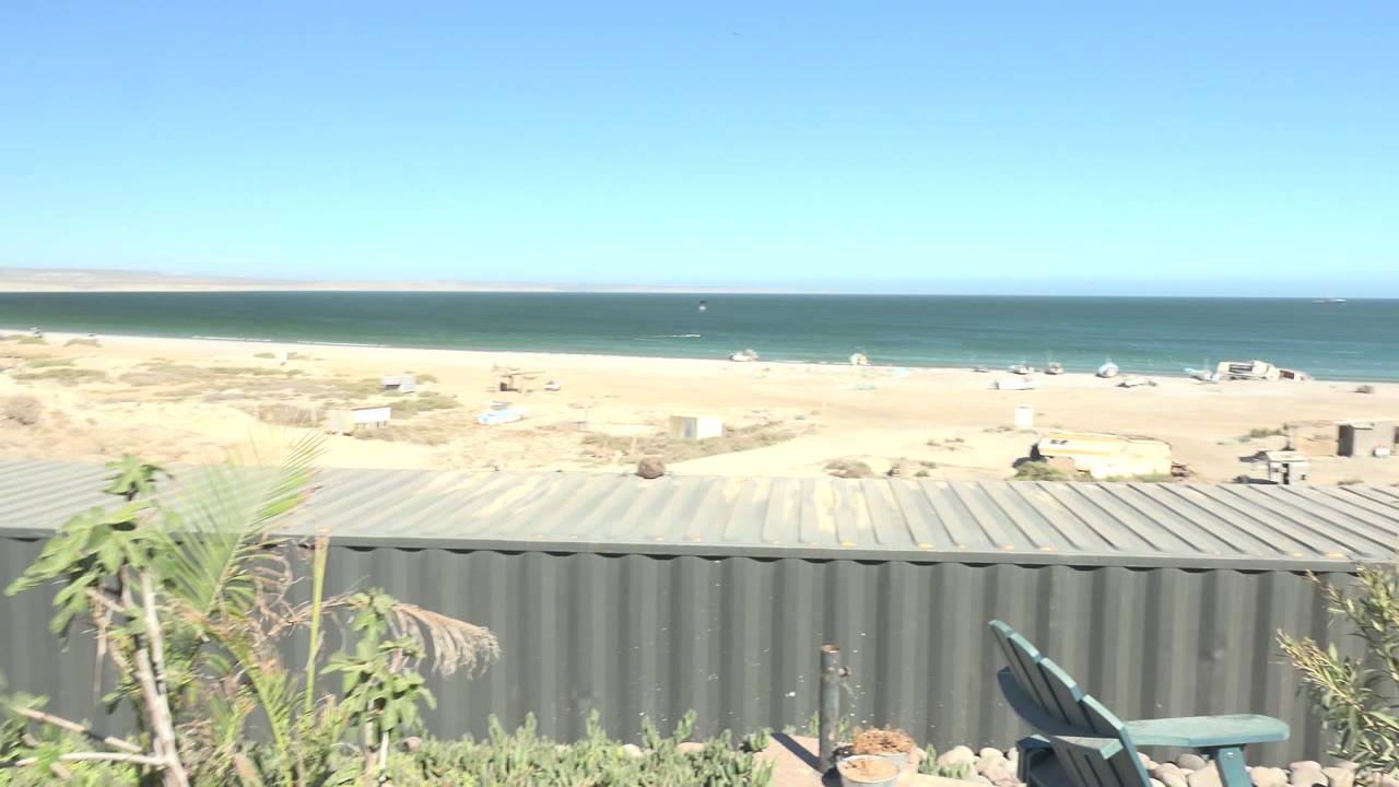 A bright elevated San Juanico coastline frame shows foreground vegetation, gray metal roofing or wall, a wide sandy beach, calm ocean, clear sky, and no visible people.