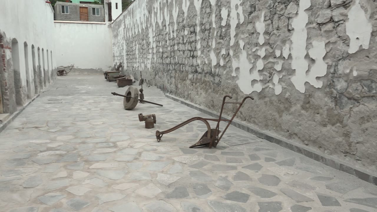 Opening frame of a Mulege, Baja California Sur stone courtyard path, with a peeling white wall on the right, arched white building on the left, rusted metal tools on the paving, and no visible people.