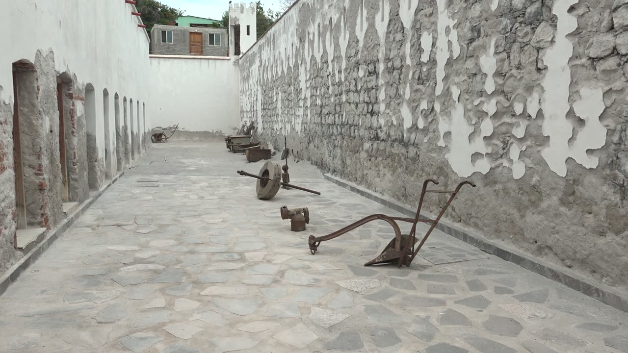 The Mulege passage remains centered as rusted metal tools sit on pale stone paving, the peeling wall texture fills the right side, and the white arched building lines the left with no visible people.