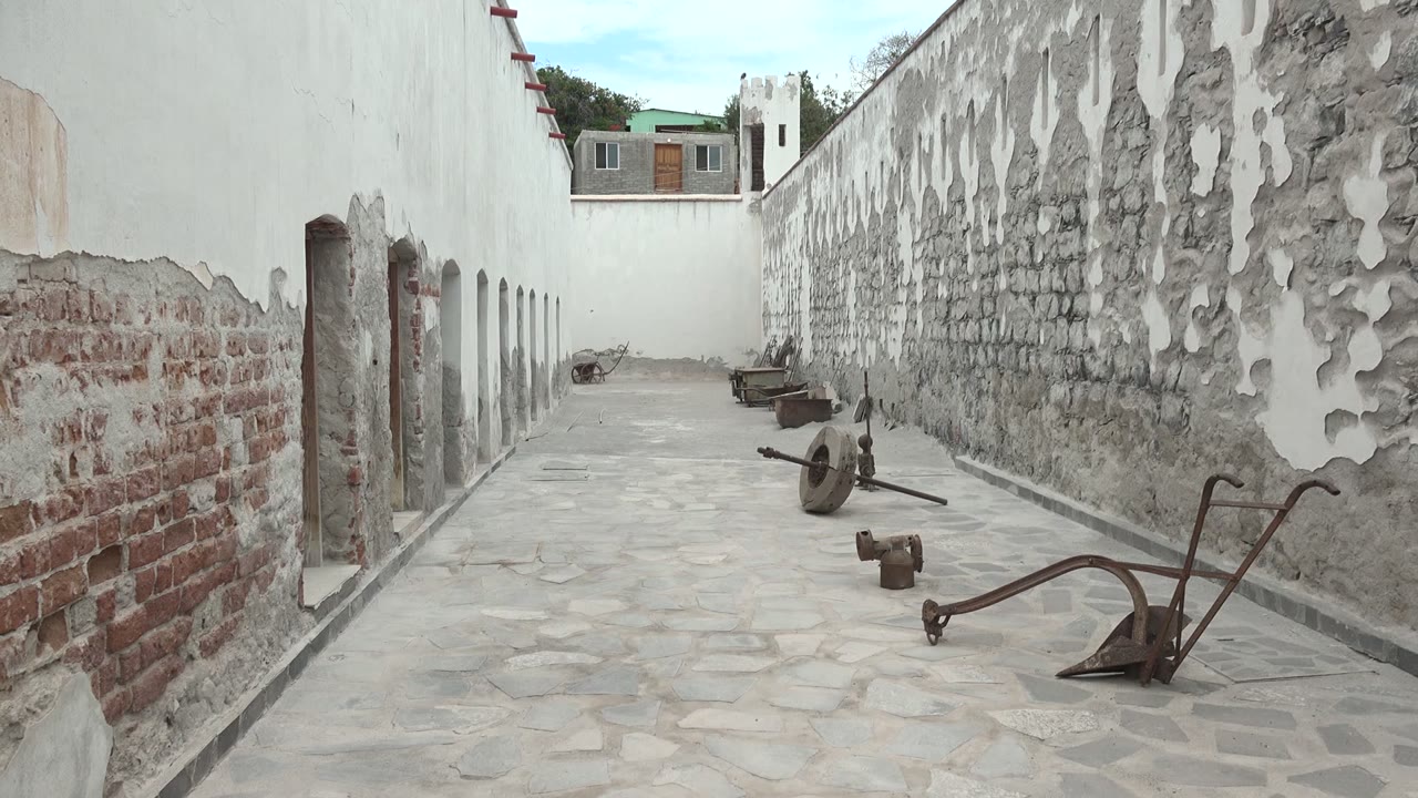 Narrow Mulege courtyard with exposed brick, peeling white plaster, cobblestone paving, rusted tools on the ground, and no visible people at 0:00.