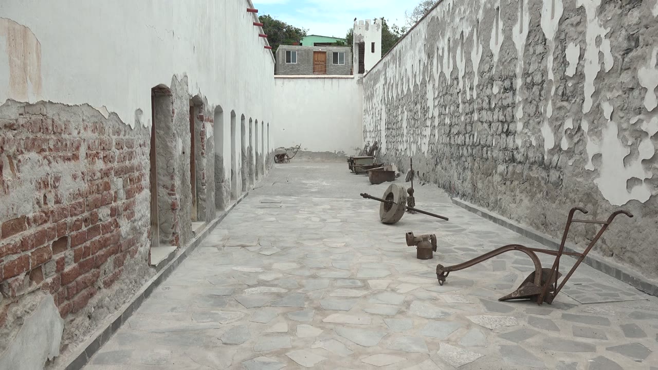 Narrow Mulege courtyard seen from ground level, with red brick exposed beneath flaking plaster, old tools resting along the path, and no visible people at 0:02.