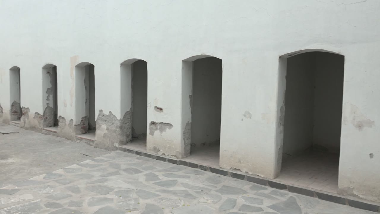 White arched doorways and worn lower plaster line a stone courtyard in Mulege, Baja California Sur, with no visible people at the opening moment.