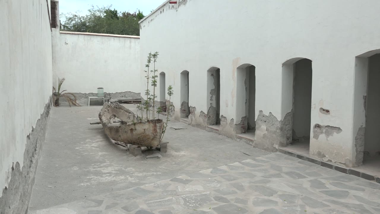 Opening frame of a Mulege courtyard with white plaster walls, stone paving, arched openings on the right, a boat-shaped planter in the center, and no visible people.