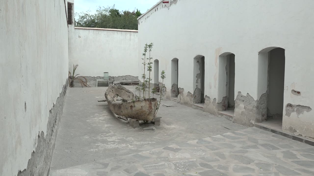 At two seconds, the Mulege courtyard remains empty, with a boat-shaped planter near center, pale stone paving below, white walls on both sides, and arched openings receding to the right.