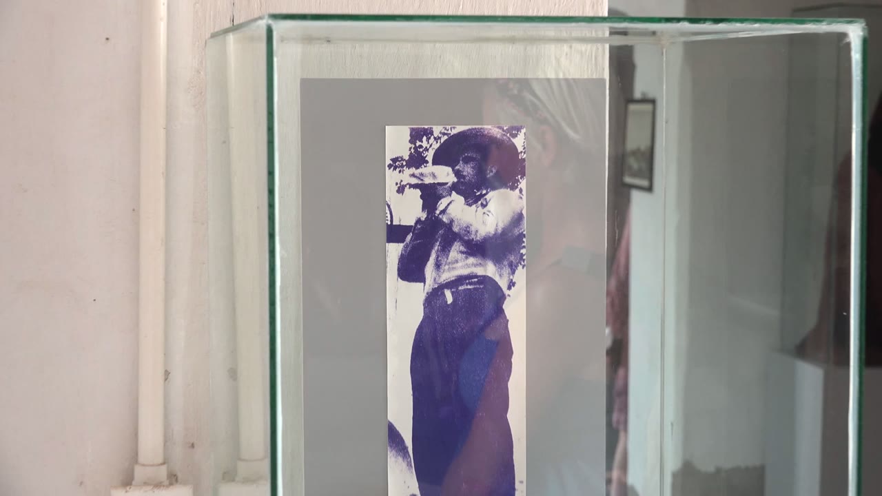 Opening frame in Mulege, Baja California Sur shows a glass display case with a small black-and-white photograph of a hat-wearing man drinking from a cup, with a white wall and vertical pipe behind it.