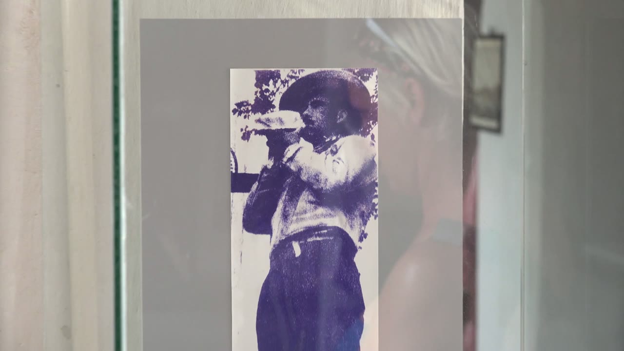 At 0:02, the photograph of the man drinking from a cup occupies more of the frame, with the case edge, gray mount, and pale wall still visible around it in Mulege.
