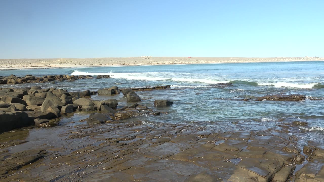 At La Bocana, Baja California Sur, dark shoreline rocks fill the foreground while low waves break offshore beneath a clear blue sky, with no visible people.