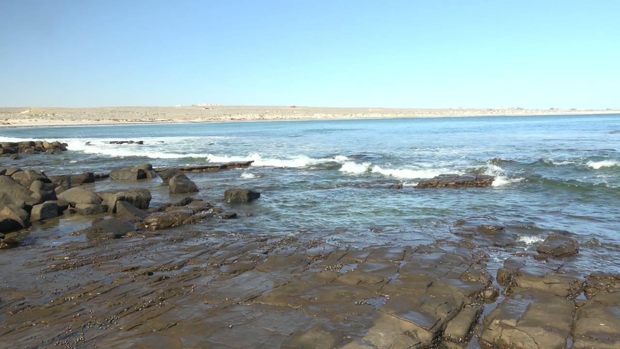 At La Bocana, Baja California Sur, the water spreads across flat foreground rocks while broken white surf sits midframe and the pale landmass runs along the horizon, with no visible people.