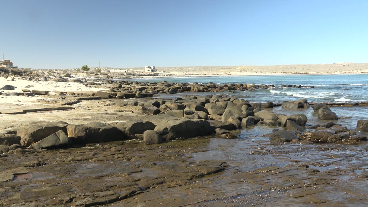 La Bocana rocky shore at the start of the clip, with wet flat rocks in front, dark boulders near the water, small distant structures on the pale horizon, and no visible people.