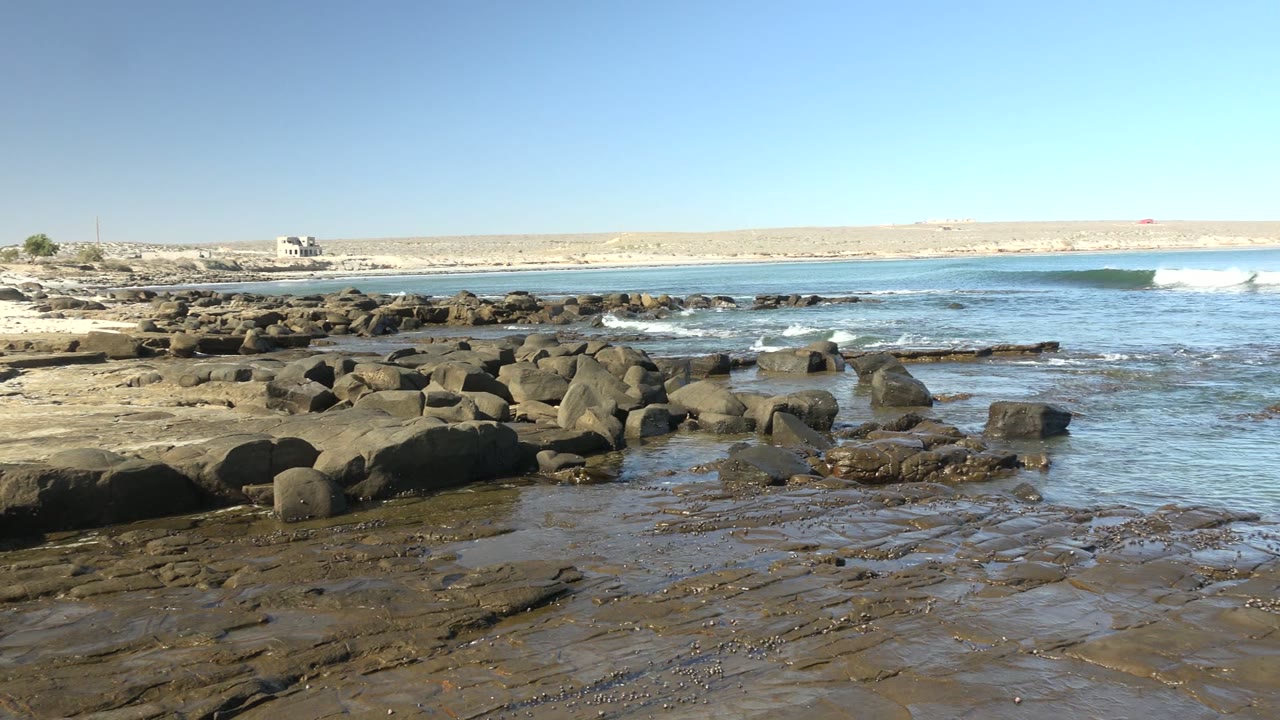 La Bocana rocky beach with a small wave crest forming to the right of the frame, wet stone ledges in front, distant pale land across the horizon, and no visible people.