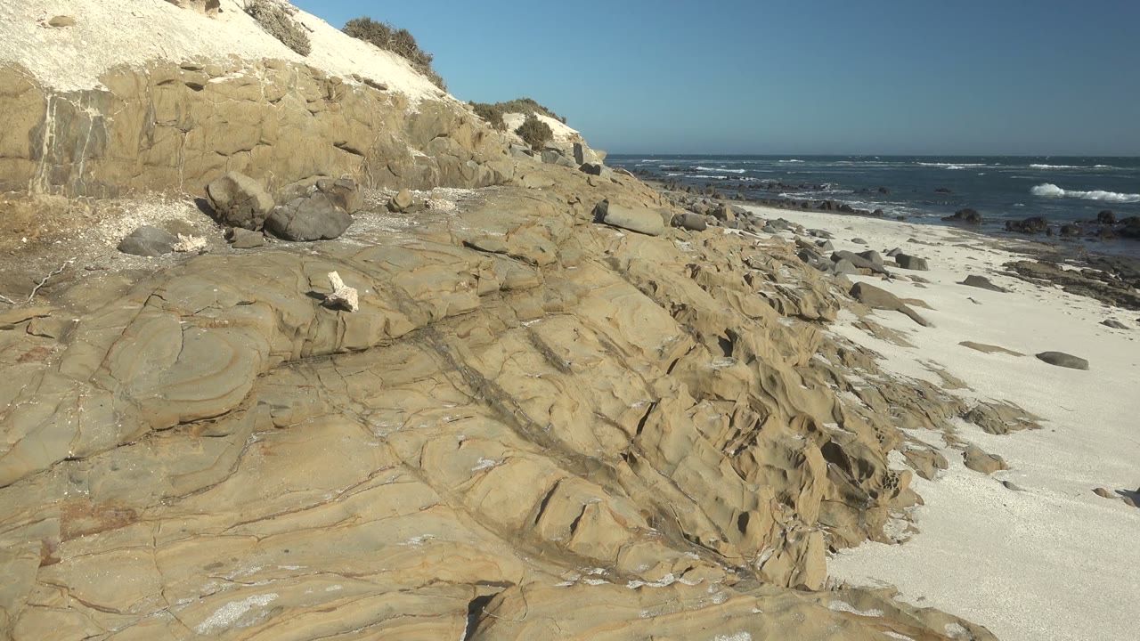 Layered tan sandstone and scattered rocks border a white sandy beach in La Bocana, Baja California Sur, with dark blue surf beyond and no visible people.