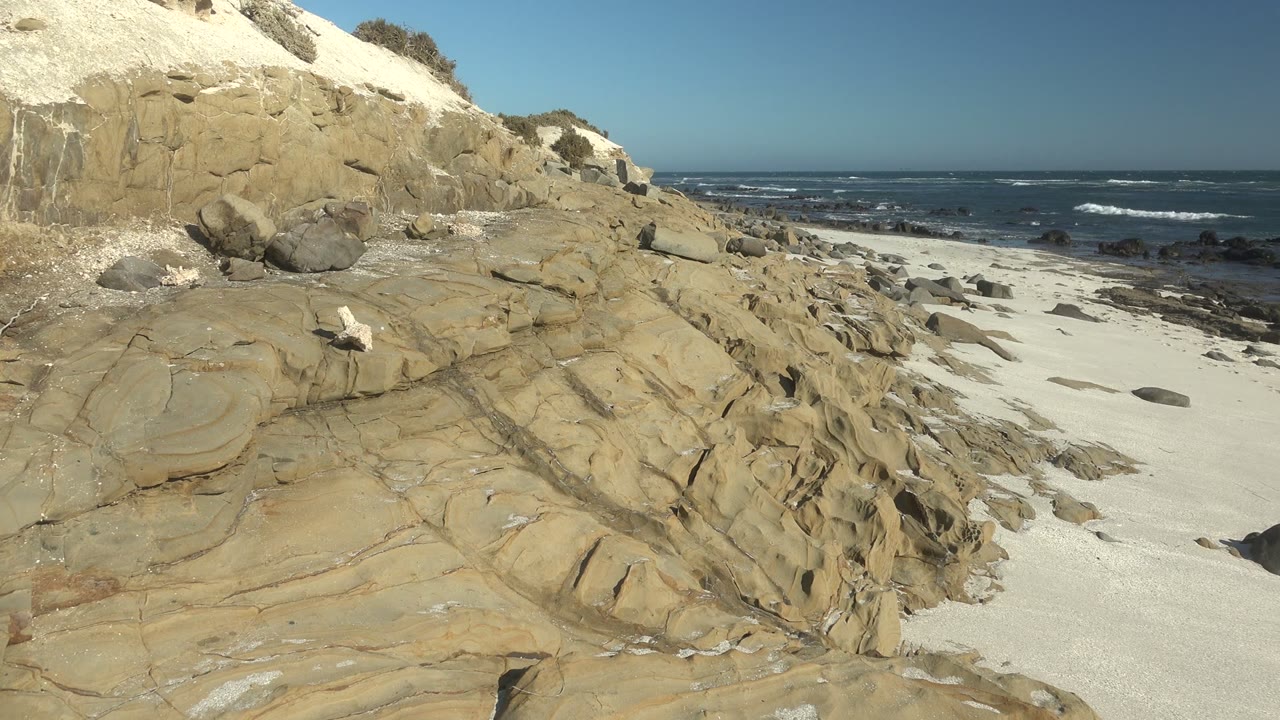 Tan coastal rocks leading toward surf and white sand.