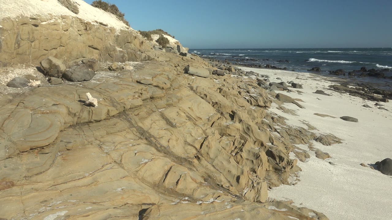 A layered cliff face and sloped sandstone sit beside pale sand in La Bocana, while waves break across the dark ocean horizon and no visible people appear.