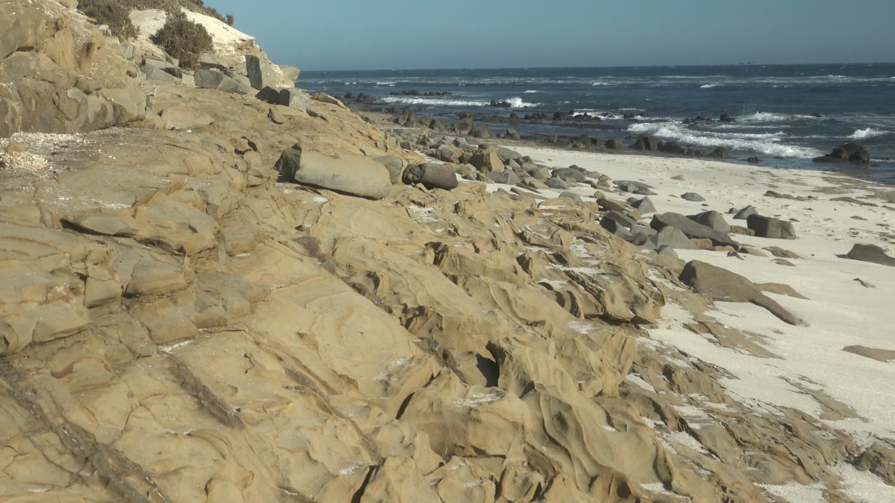 Opening view of tan rock shelves and scattered boulders on the La Bocana shore, with white sand leading to dark blue surf and no visible people.