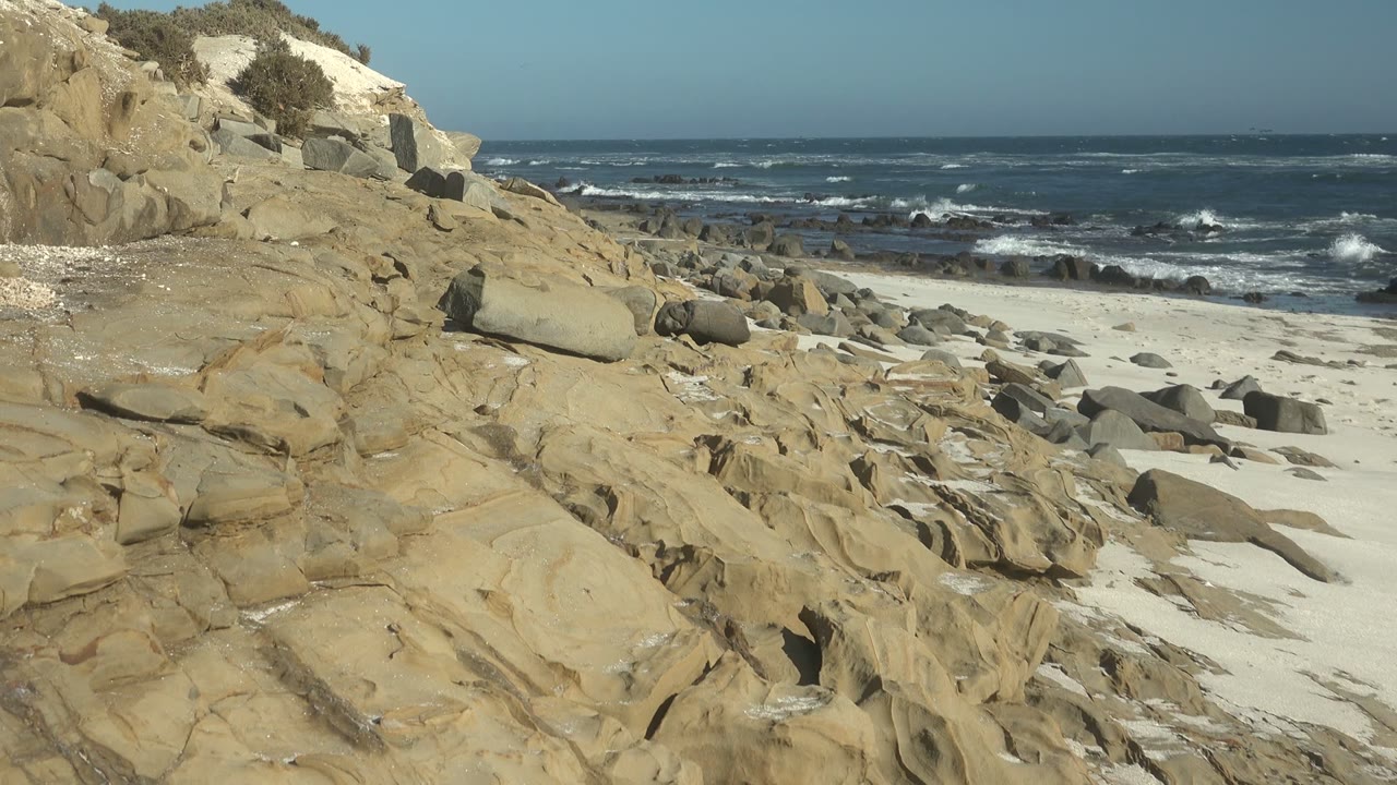 Sunlit coastal rocks, white sand, and breaking waves.