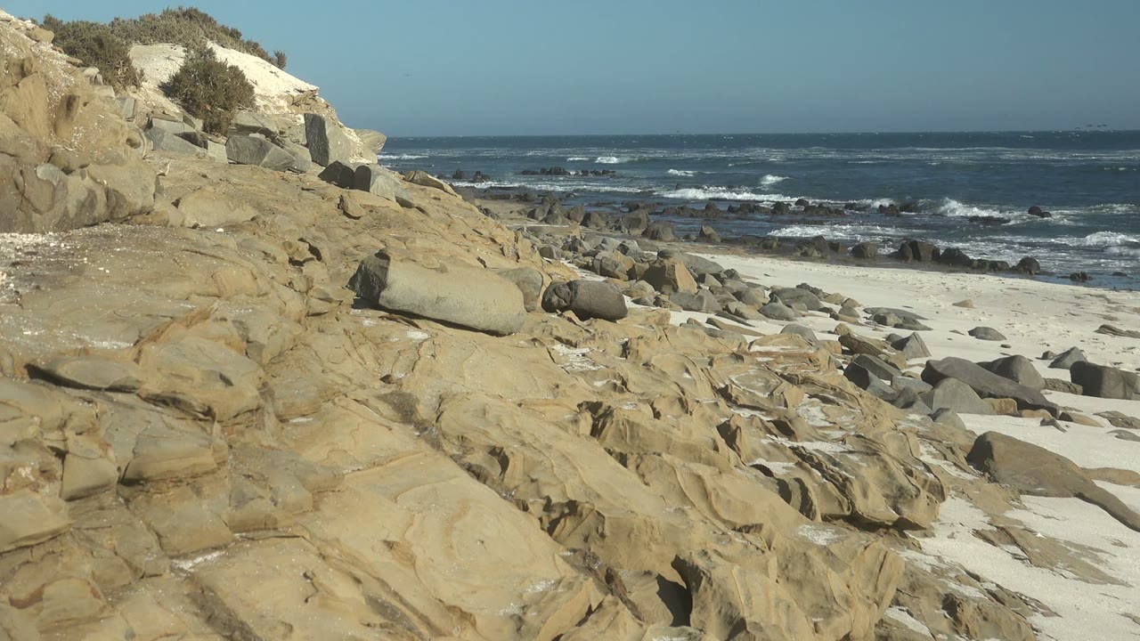 A pale rocky slope with shrubs rises at the left of the La Bocana beach while boulders, sand, and whitecaps stretch across the frame, no visible people.