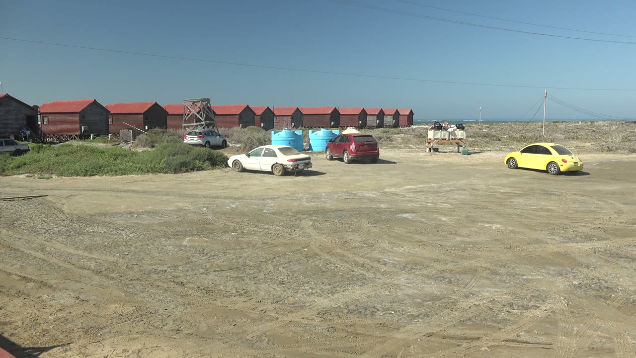 Opening view in La Bocana, Baja California Sur shows a wide dirt lot, red-roofed buildings, blue containers, parked white, red, and yellow cars, and no visible people.