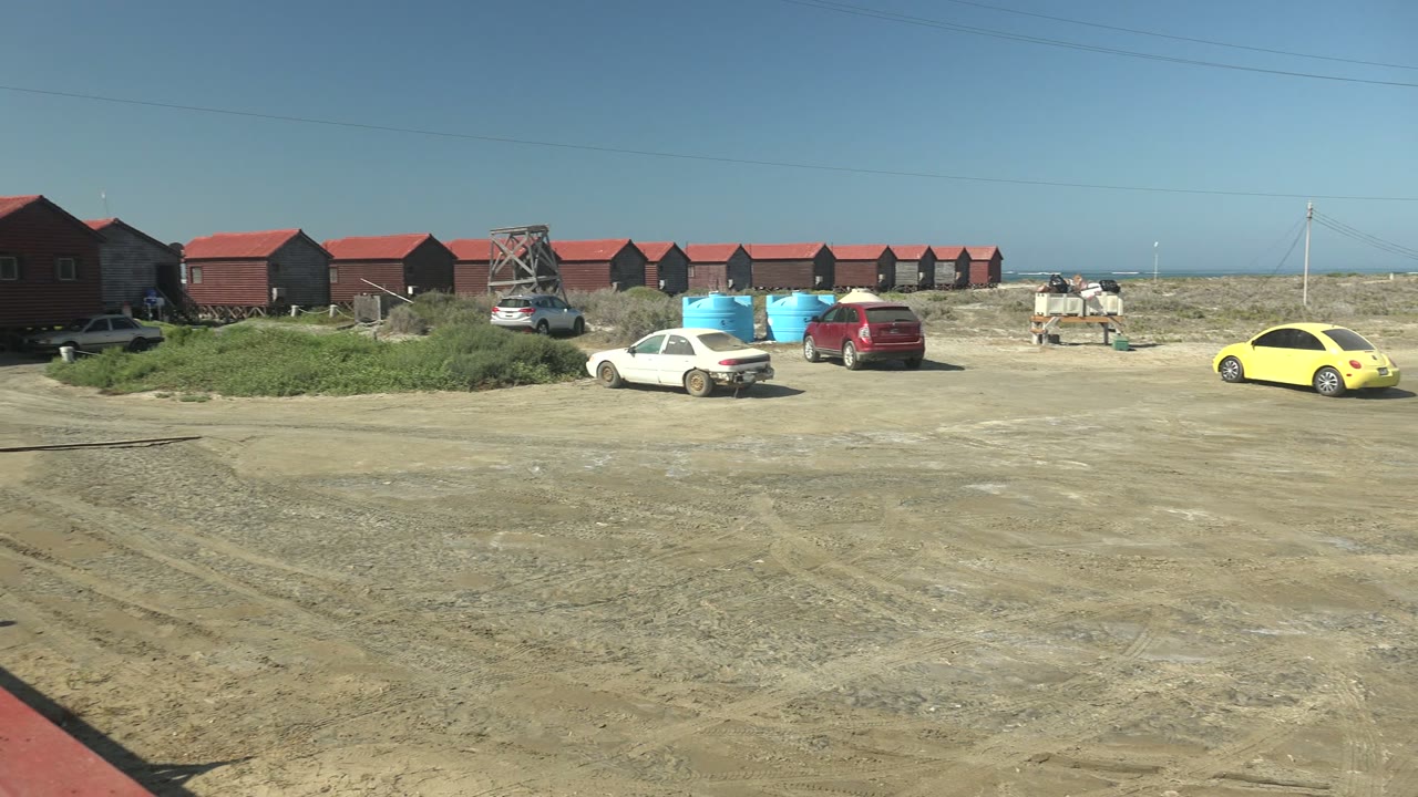 A slightly shifted La Bocana view shows the same dirt lot, red-roofed structures, blue containers, utility lines, and parked cars with no visible people.