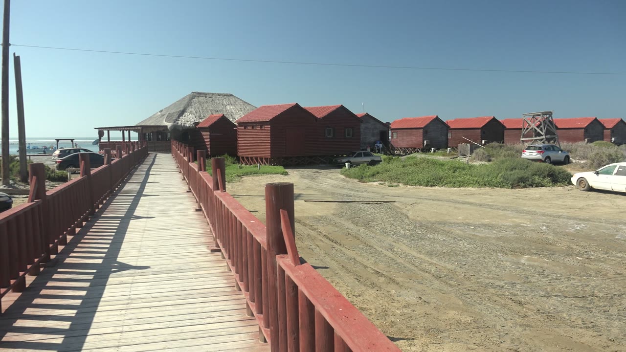 A wooden boardwalk with red railings leads toward red-roofed buildings in La Bocana, Baja California Sur, with gravel parking, ocean horizon, clear sky, and no visible people.