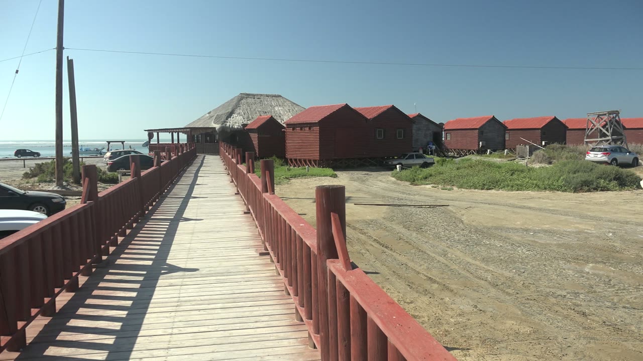 Boardwalk and red buildings beside a gravel lot with vehicles near the ocean.