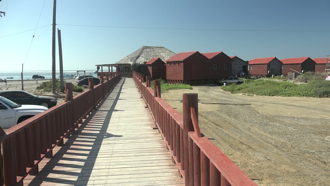 A red-railed boardwalk in La Bocana points toward clustered red buildings, with a white vehicle at the lower right, gravel road surface, ocean horizon, and no visible people.