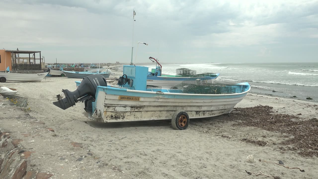 At Punta Abreojos, Baja California Sur, a white and blue fishing boat with an outboard motor rests on sand while waves break in the background and no people are visible.