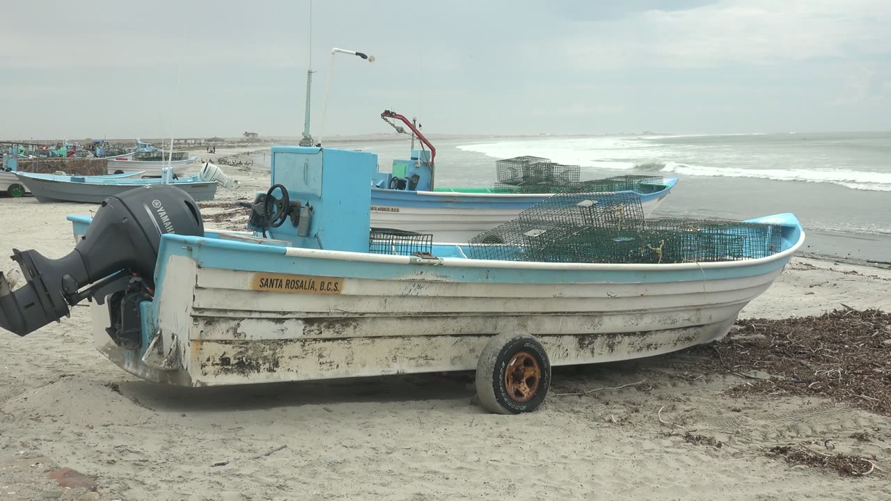 At 0:00, a white and blue fishing boat fills the foreground on the sandy beach at Punta Abreojos, Baja California Sur, with ocean surf and gray sky behind it and no visible people.