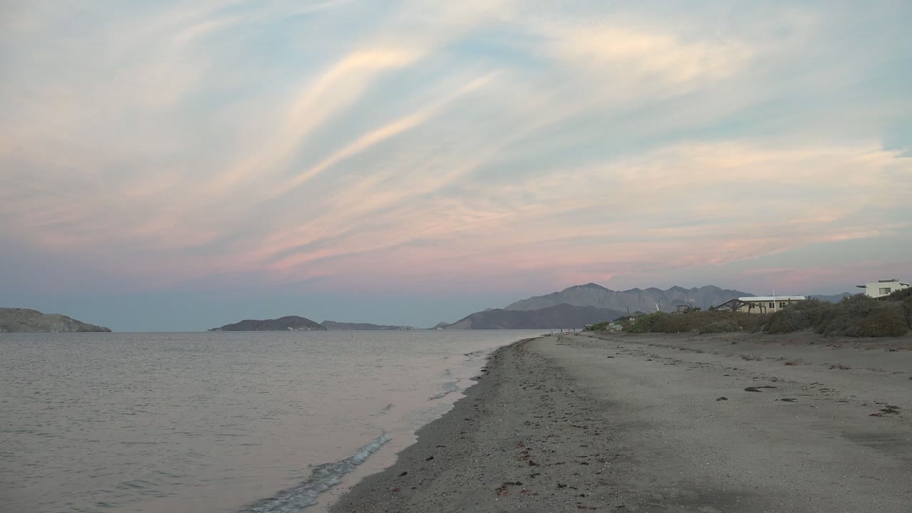 Bahia De Los Angeles sandy shoreline at dusk with gentle water on the left, a narrow beach curve, distant landforms, pastel clouds, and no visible people.