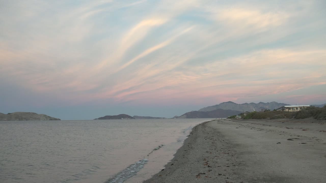 A Bahia De Los Angeles beach curve runs beside gently rippled water toward low distant mountains beneath streaked pastel clouds, with no visible people.