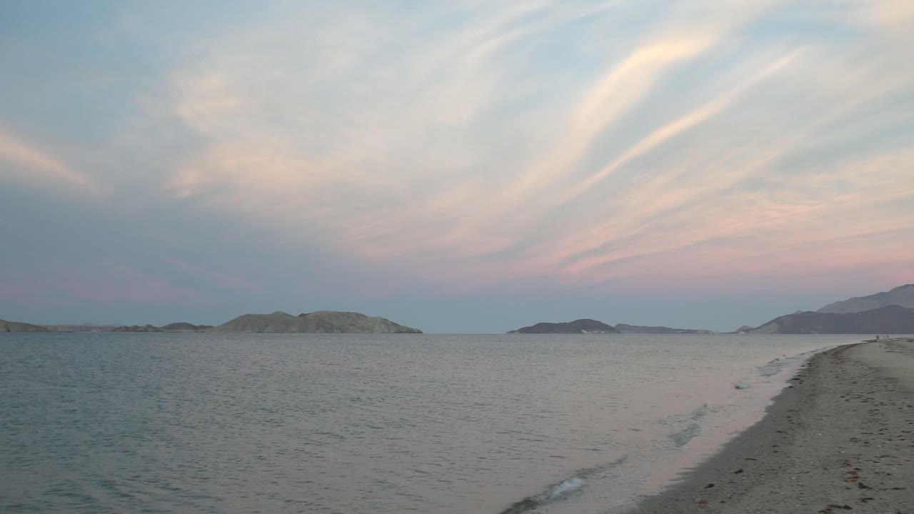 Opening view of Bahia De Los Angeles with pale sand in the foreground, calm ocean, low islands on the horizon, pink-streaked clouds above, and no visible people.