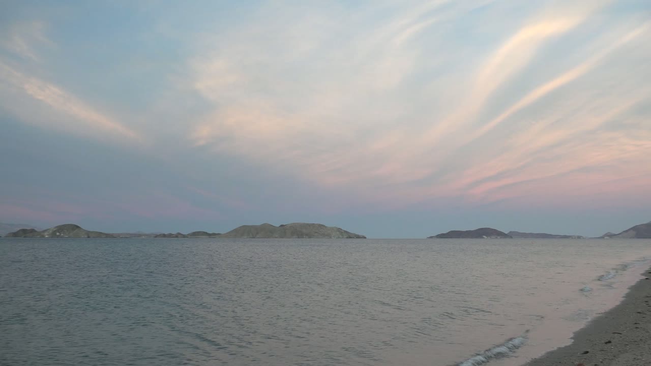 Two seconds into the Bahia De Los Angeles pan, a narrow sandy shoreline curves through the foreground toward calm water, distant islands, and no visible people.