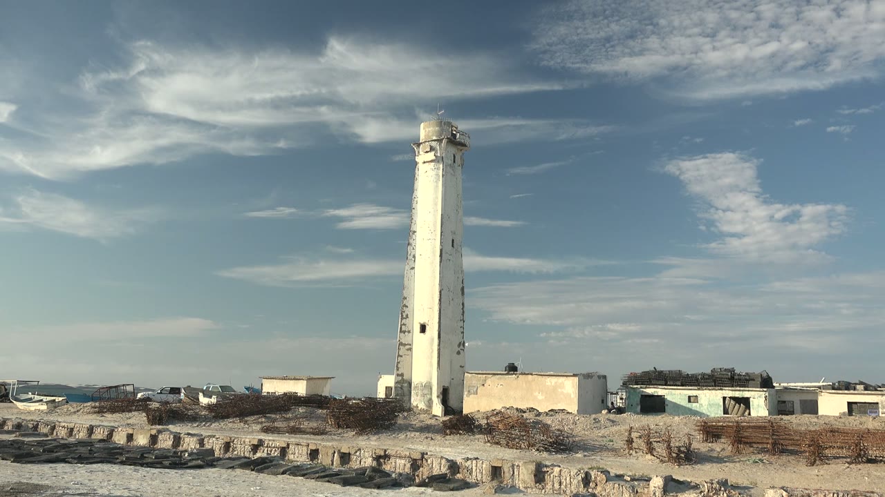 A white lighthouse stands on the Guerrero Negro sandy coast with low buildings and wispy clouds overhead, with no visible people.